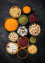 Legumes, lentils, chikpea and beans assortment in different bowls on black stone table. Top view.