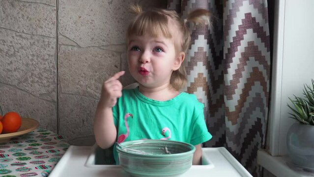 A little girl in a green dress sits at a high chair and eats baby porridge from the table with her fingers and looks out the window.child eating porridge with fingers