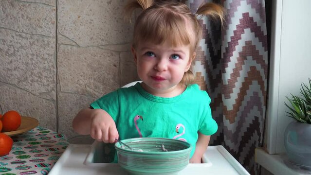 A little girl in a green dress sits at a high chair and eats baby porridge from the table with her fingers and looks out the window.child eating porridge with fingers