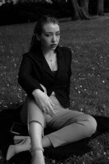 Black and white portrait of a young girl in the city park. Elegant and melancholy portrait, the girl is sitting on the grass. Beautiful woman in classic clothes