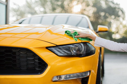 Closeup cropped image of hand of young confident stylish european businesswoman washing and cleaning yellow sport car with microfiber cloth, wiping hood and headlights. Car wash outdoors concept.