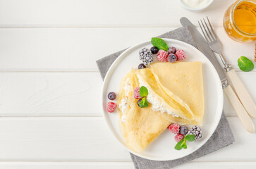 Traditional thin pancakes or crepes with cottage cheese and raisins with berries, honey and sour cream on a white wooden background. Food for Maslenitsa. Copy space.