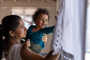 Happy Latin American mom (30) and her son (2) is lifted in arms and look out the window while playing at home. Mother's Day concept, single mother.