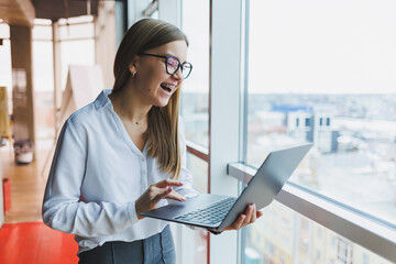 Cheerful young fair-haired business woman of European appearance in a white shirt and glasses holds a portable laptop in her hands, stands near a large window in the office on the top floor