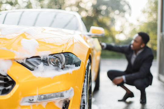 Crop close up of car headlight. Horizontal image of African businessman in formal wear washing his yellow luxury car using yellow sponge for cleaning car door with soap foam