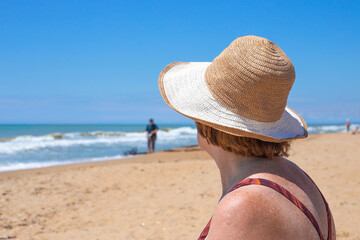 Mature woman in a hat on the sandy shore looks at the sea, side view. Rest of pensioners in Russia