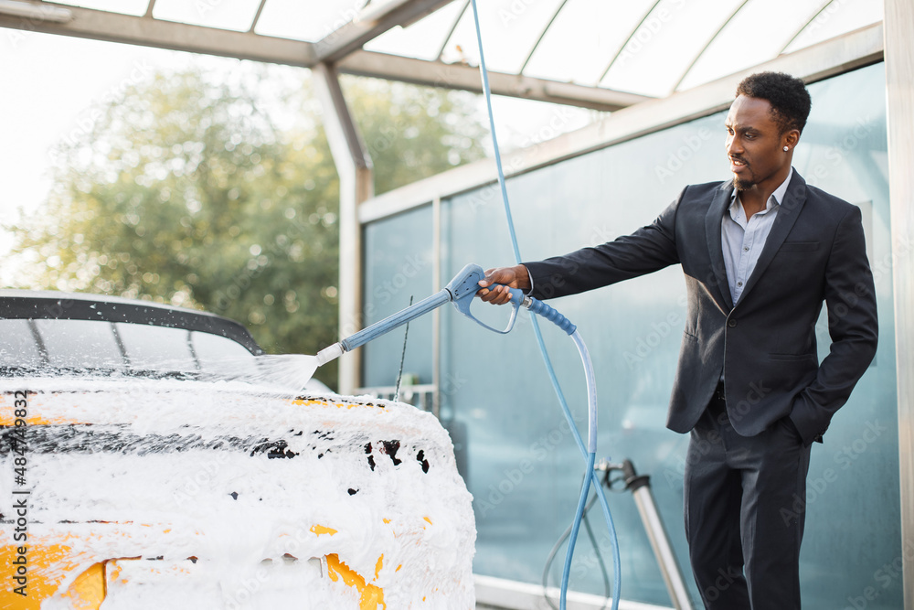 Poster Portrait of handsome bearded young African businessman washing his yellow car with foam at self service carwash outdoors. Clean car concept. Car wash.