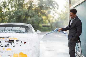 Serious african man in business suit, at self car wash outdoors, is spraying cleaning foam to a modern yellow sport luxury car holding a high pressure