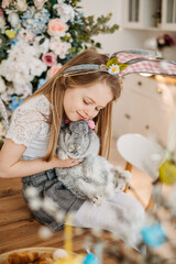 A cute little girl in a wooden kitchen at home hugs a gray rabbit in her arms. Easter theme