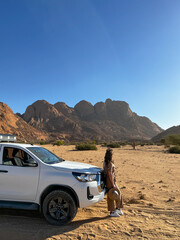 Obraz premium Young woman in overalls with hat stands near car in desert. Spitzkoppe mountain.