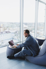 Break. Smiling young man relaxing on bean bag chair while sitting at table and relaxing using laptop, happy businessman leaning back, enjoying work, feeling satisfied