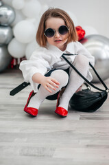A fashionable baby in sunglasses is trying on her mom's red shoes. Fashion and modern clothes....