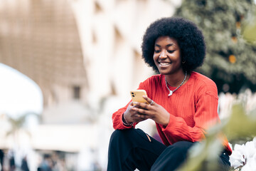Smiling young black woman with afro hairstyle using her smart phone on the street