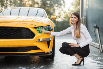 Cheerful business woman washing and wiping modern sport yellow car headlight at the outdoor car...