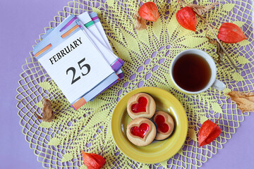 Fototapeta na wymiar Calendar for February 25: the name of the month February in English, the numbers 25 on the loose-leaf calendar, a cup of tea, heart-shaped cookies, physalis branches on a yellow openwork napkin