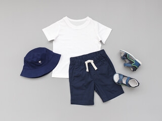 White t-shirt, blue shorts, sandals and panama hat on grey background. Children's clothing. Summer outfit for a boy. Flat lay, top view.