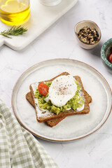 Trendy lifestyle sandwich: protein bread slice with cream cheese, mashed avocado, cherry tomatoes and poached egg on white scandi plate, light setting