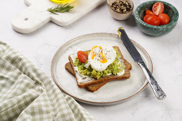 Trendy lifestyle sandwich: protein bread slice with cream cheese, mashed avocado, cherry tomatoes and poached egg on white scandi plate, light setting
