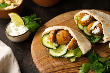 Traditional oriental chickpea deep fried falafel in flatbread pita, tzatziki yoghurt sauce, hummus, fresh lime and green cilantro on wooden board, dark surface