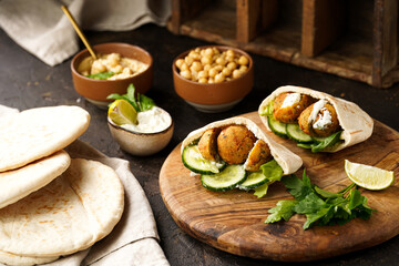 Traditional oriental chickpea deep fried falafel in flatbread pita, tzatziki yoghurt sauce, hummus, fresh lime and green cilantro on wooden board, dark surface