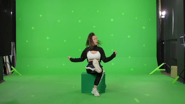 A young woman dancing on a green screen background. Attractive girl making content for social media. Female danser sits on box. Chroma key