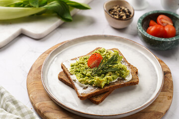 Trendy lifestyle sandwich: protein bread slice with cream cheese, mashed avocado, cherry tomatoes and rosemary on white scandi plate, light setting