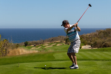 Middle aged man golfing, caught mid swing off the tee box. On ocean side tropical golf course with...