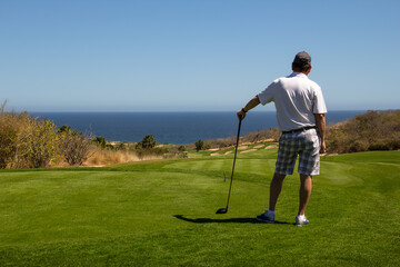 Middle aged man wearing golf shorts and golf shirt, looking out over ocean side tropical golf...