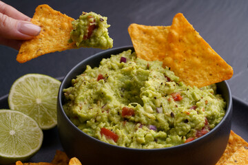 Closeup of woman hand with tortilla chips or nachos with fresh tasty guacamole dip in black plate...