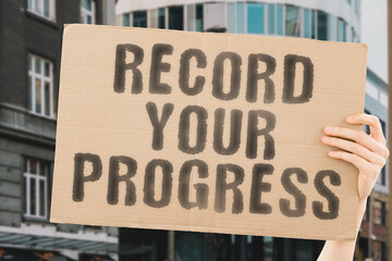 The phrase " Record your progress " on a banner in men's hand with blurred background. Rise. Increase. Improve. Evolution. Achieved. Level up. Updated. Updating. Tracking. Working. Worker. Time