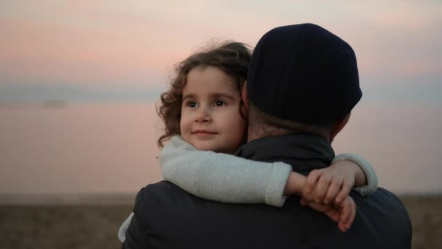 Happy adorable little girl with curly hair hugs her father and smiles. Portrait of a girl with her father on the beach. Father and daughter love and comfort support concept