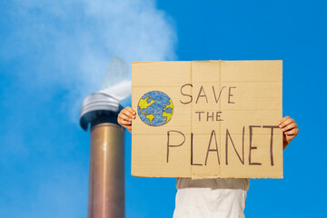 an unrecognizable young woman holds a sign with the text "save the planet" in front of a smoky chimney.