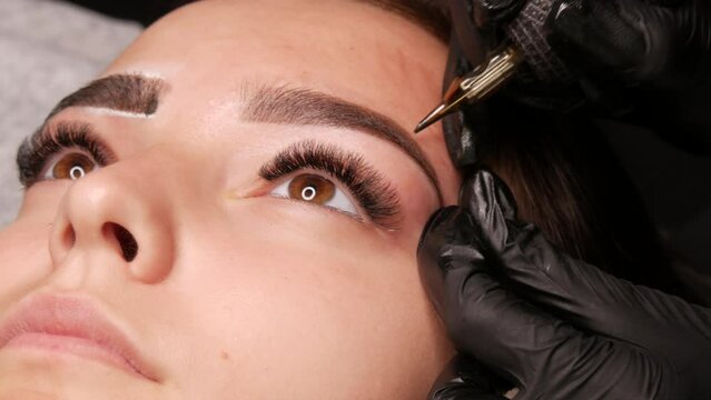 Eyebrow permanent make-up. The master in the studio introduces black pigment under the skin of a young model using a special machine. Modern tattoo for beauty