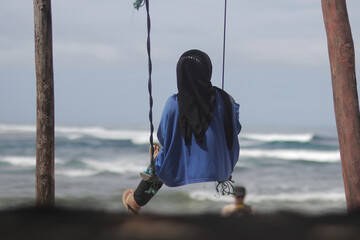 A young Asian woman in a black hijab and blue shirt is sitting on the swing near the Cibuaya beach Ujung Genteng, Sukabumi, West Java, Indonesia.