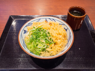 closed-up tanuki udon with chives and crunchy bits of deep-fried dough produced as a byproduct of cooking tempura and cup of japanese tea on the black tray