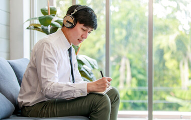 Asian young handsome professional successful male businessman employee in formal business shirt and necktie sitting on cozy sofa listening to streaming music online browsing internet .