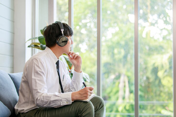 Asian young handsome professional successful male businessman employee in formal business shirt and necktie sitting on cozy sofa listening to streaming music online browsing internet .