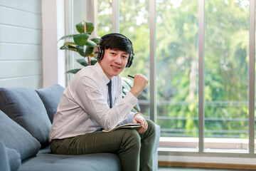 Asian young handsome professional successful male businessman employee in formal business shirt and necktie sitting on cozy sofa listening to streaming music online browsing internet .