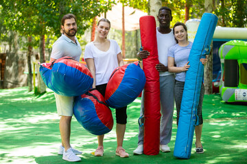Fototapeta na wymiar Portrait of cheerful multiracial group of adult people standing with inflatable boxing gloves and logs in outdoor amusement park