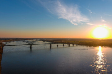 Fototapeta na wymiar the gorgeous vast flowing water of the Mississippi river with a stunning blue, yellow and red sunset in the sky with a bridge over the water and vast trees along the banks at Green Belt Park