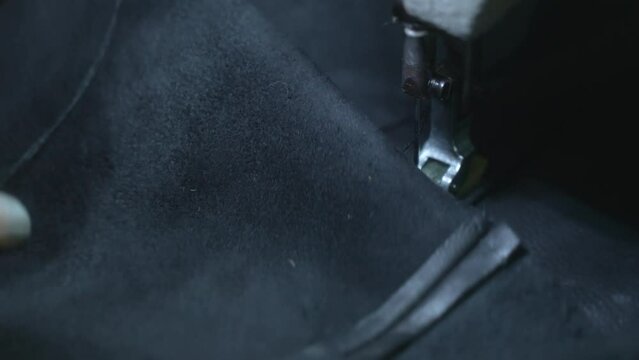Close-up of sewing a needle seam on a black leather jacket on a sewing machine. Seamstress creates leather goods, stitches a line in a dark factory room.