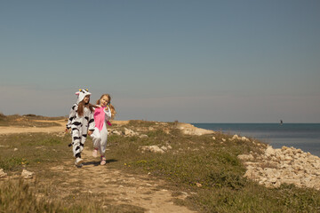 Two pretty girls in kigurumi cow and unicorn walk on the seashore, friends chat and play