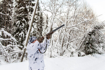 The concept of winter hunting. A male hunter in a white camouflage suit shoots a gun.