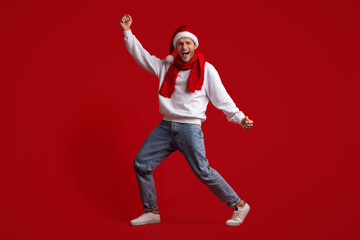 Fototapeta na wymiar Xmas Joy. Portrait Of Dancing Millennial Guy Wearing Santa Hat And Scarf