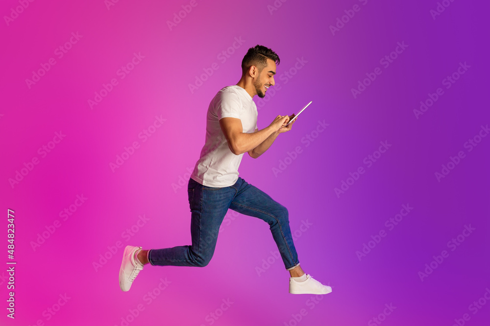 Poster full length of young arab guy with tablet computer jumping in neon light, free space. cool app or ad