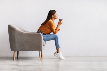 Side view of young Asian woman sitting in armchair, drinking hot coffee, relaxing against white wall, free space