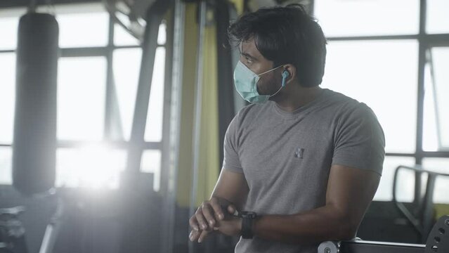 Young muscular builder with medical face mask checking workout plan or monitoring hart rate from smartwatch at gym - concpet of healthcare, technology and safety measures during covid-19 pandemic.
