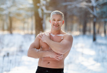 Positive senior man tempering his body with snow at frosty winter forest. Cold exposure training