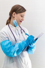 Beautiful young woman doctor in a medical gown, mask and blue gloves with a stethoscope writes down a medical history in a hospital. Selective focus. Portrait. Close-up