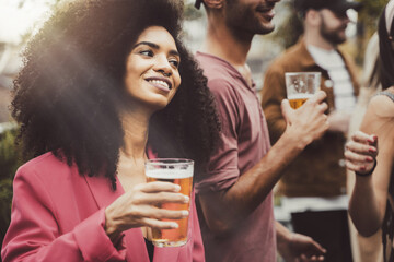 cheerful trendy Brazilian girl in a students rooftop party holding a glass of beer - Group of people having fun drinking and dancing in the terrace